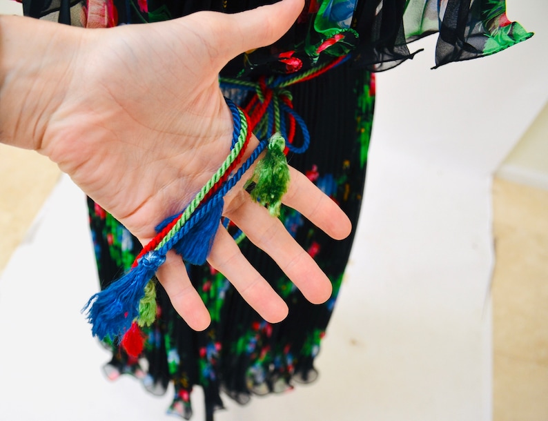 May include: Black pleated maxi dress with a floral print and colorful tassel ties. The dress has a fitted bodice and a flowing skirt.