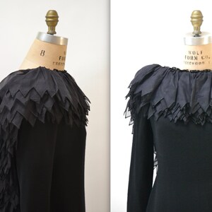 May include: A black long-sleeved dress with a large, layered ruffle collar. The ruffle is made of sheer fabric and has a scalloped edge.