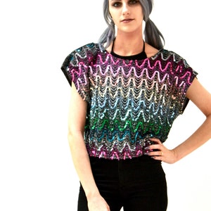 Puede incluir: Una blusa de lentejuelas arcoíris con un patrón ondulado. La blusa tiene mangas cortas y un escote redondo.