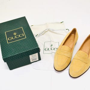 May include: A pair of yellow suede Gucci loafers with a gold logo on the dust bag. The loafers have a classic loafer design with a tassel and a leather sole.