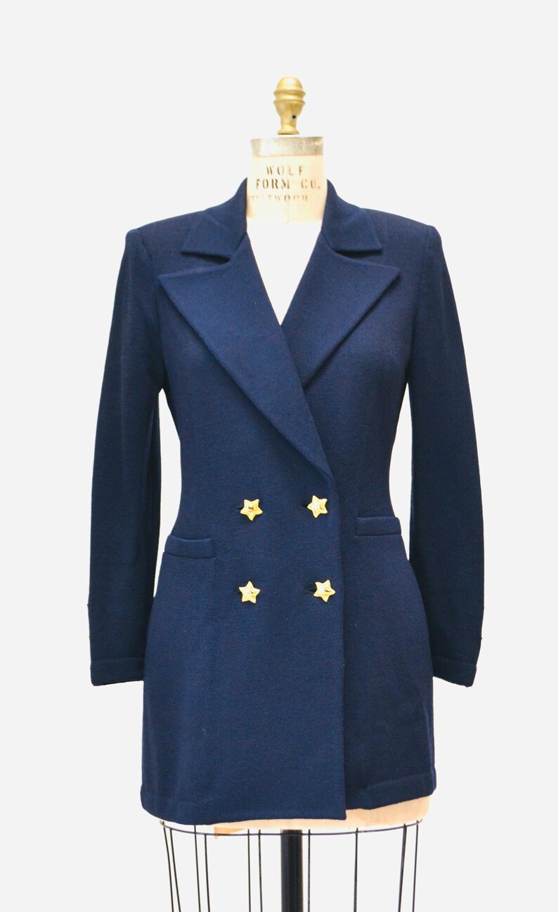 May include: A navy blue blazer with a double breasted closure and gold star buttons. The blazer has a long sleeve and a fitted silhouette.