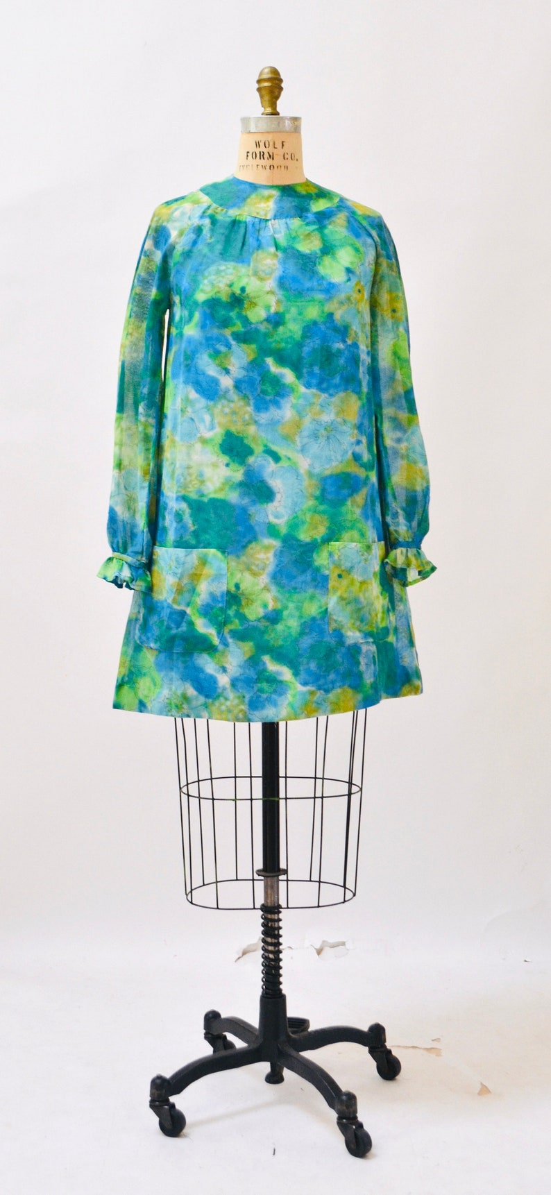 May include: A blue and green tie-dye dress with a round neckline and long sleeves. The dress is on a dress form with a black metal stand.