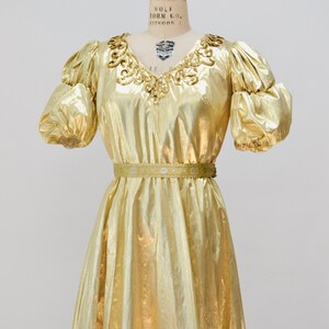 May include: A gold metallic dress with a sequined neckline and a matching belt. The dress has puffed sleeves and a flowing skirt.