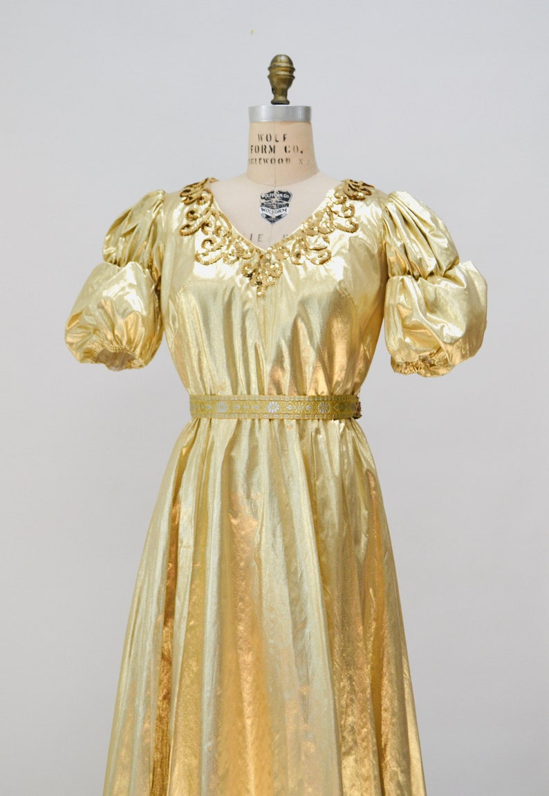 May include: A gold metallic dress with a sequined neckline and a matching belt. The dress has puffed sleeves and a flowing skirt.