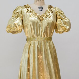 May include: A gold metallic dress with a beaded neckline and a matching belt. The dress has a flowing skirt and puffed sleeves.
