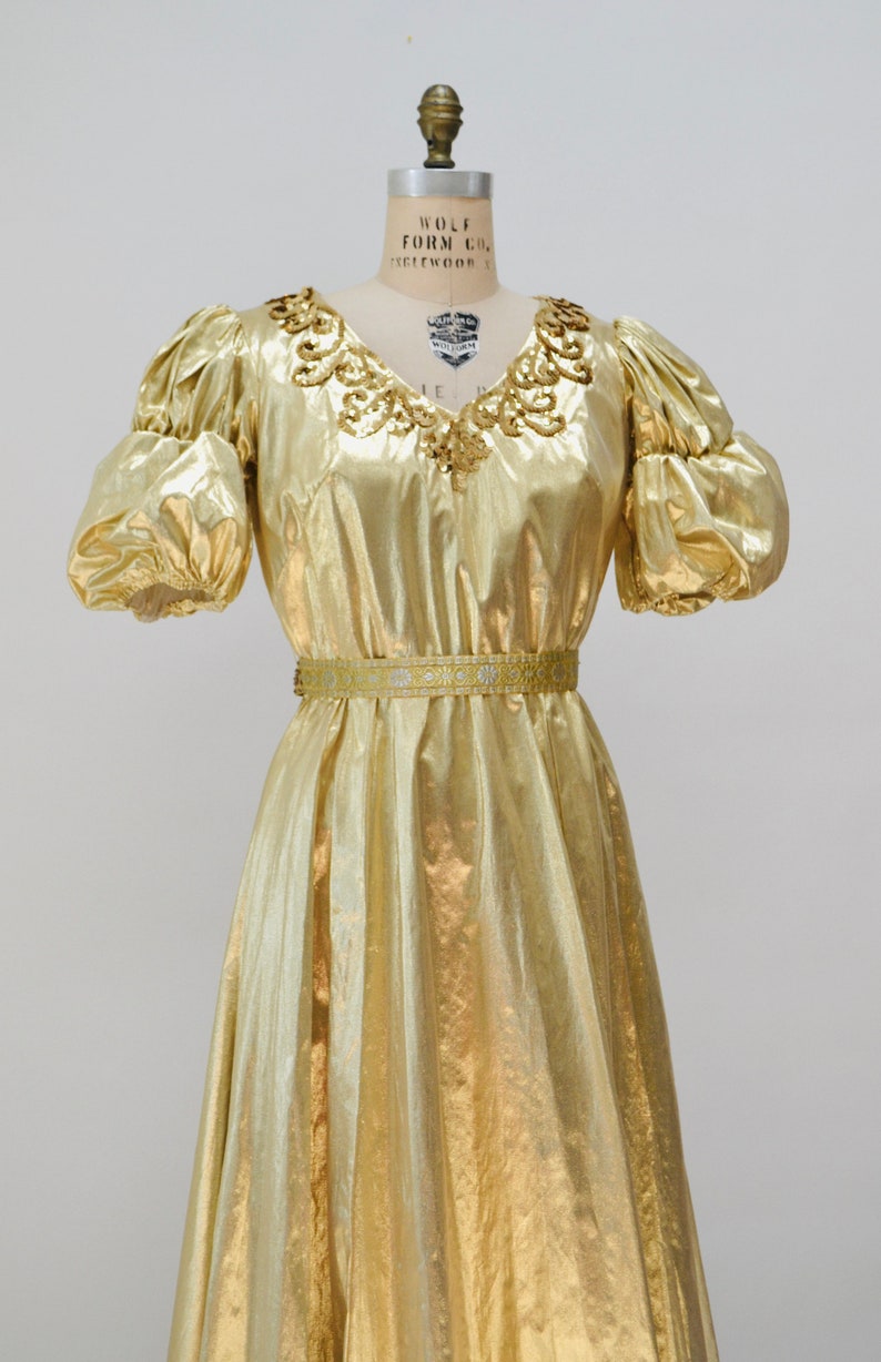 May include: A gold metallic dress with a beaded neckline and a matching belt. The dress has a flowing skirt and puffed sleeves.