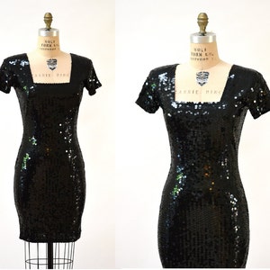 May include: A black sequined mini dress with short sleeves and a square neckline. The dress is on a mannequin.