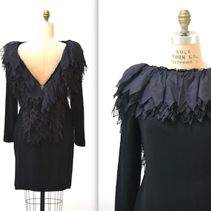 May include: A black long-sleeved dress with a ruffled neckline and a deep V-back. The ruffles are made of a sheer fabric and are layered to create a dramatic effect.