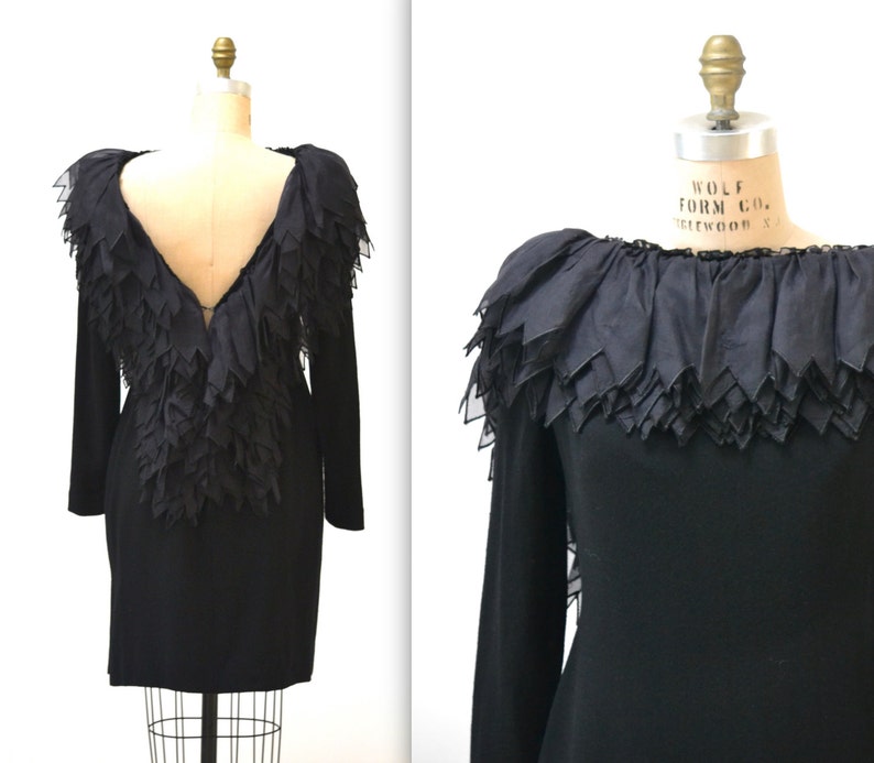 May include: A black long-sleeved dress with a ruffled neckline and a deep V-back. The ruffles are made of a sheer fabric and are layered to create a dramatic effect.