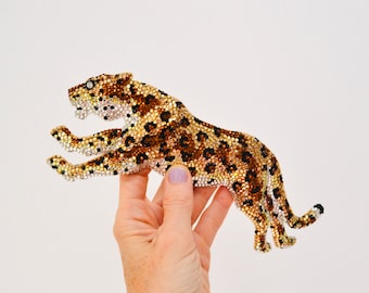 May include: A gold, brown, and black jeweled jaguar figurine. The jaguar is leaping forward with its mouth open and its tail curled behind it.
