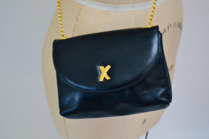 May include: A dark blue leather shoulder bag with a gold chain strap and a gold 'X' logo on the front.