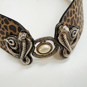Pode incluir: Um cinto com estampa de leopardo marrom e preto, com fivela de cobra prateada e centro de pérola falsa. O cinto é feito de couro e tem um design único e atraente.