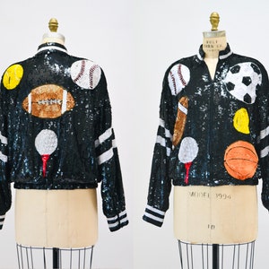 Op de afbeelding: Zwarte bomberjack met pailletten met applicaties van sportballen, waaronder een honkbal, voetbal en basketbal. De jas heeft witte strepen op de mouwen en een witte kraag. De jas wordt op een mannequin getoond.