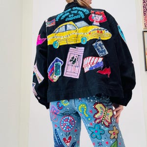 May include: Black denim jacket with colorful sequined patches, including a yellow car and text that reads "When the going gets tough, the tough go shopping." The jacket is paired with light blue jeans decorated with painted flowers, butterflies, and other whimsical designs.