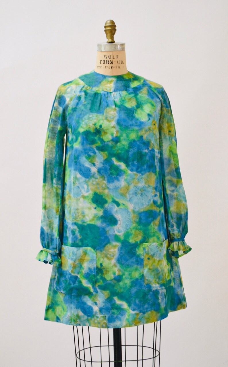 May include: A vintage 1960s tie-dye mini dress with long sleeves and a round neckline. The dress features a blue, green, and yellow tie-dye pattern. The dress has two pockets on the front and a ruffle trim on the sleeves.