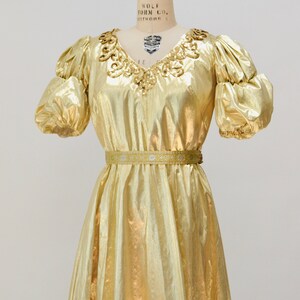 May include: A gold metallic dress with a sequined neckline and a matching belt. The dress has puffed sleeves and a flowing skirt.