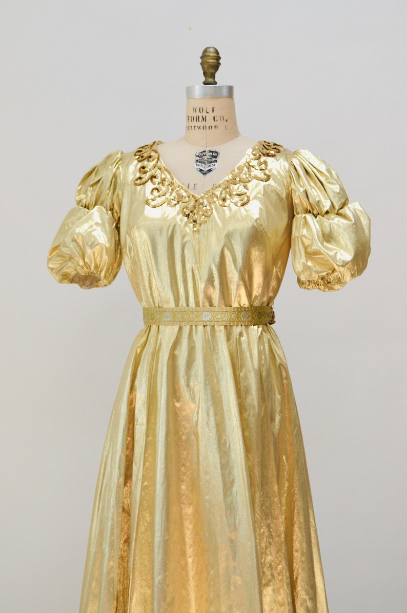 May include: A gold metallic dress with a sequined neckline and a matching belt. The dress has puffed sleeves and a flowing skirt.