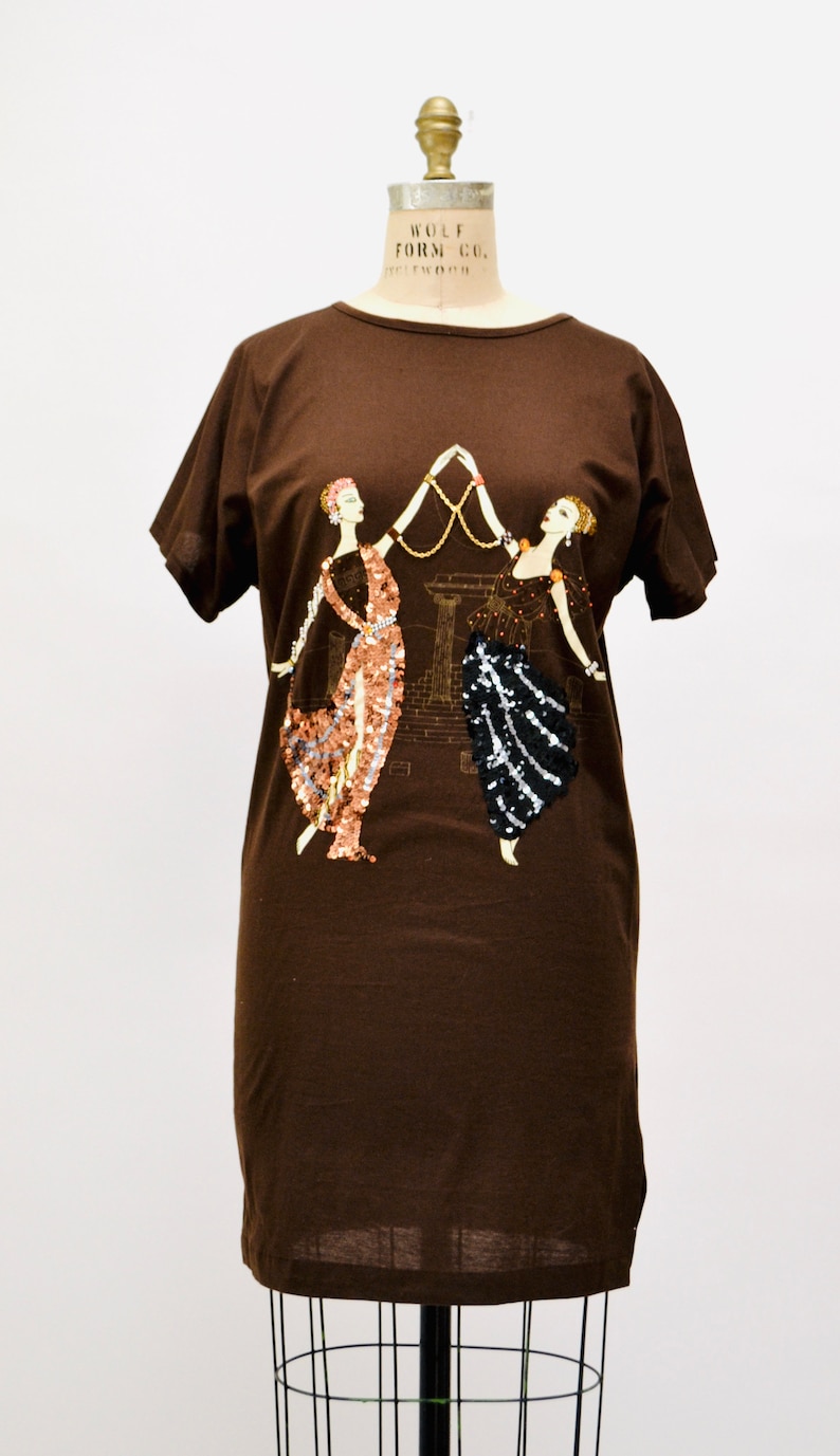 May include: A brown t-shirt dress with an art deco design of two women holding a gold necklace. One woman's dress is embellished with copper sequins, the other with black sequins.