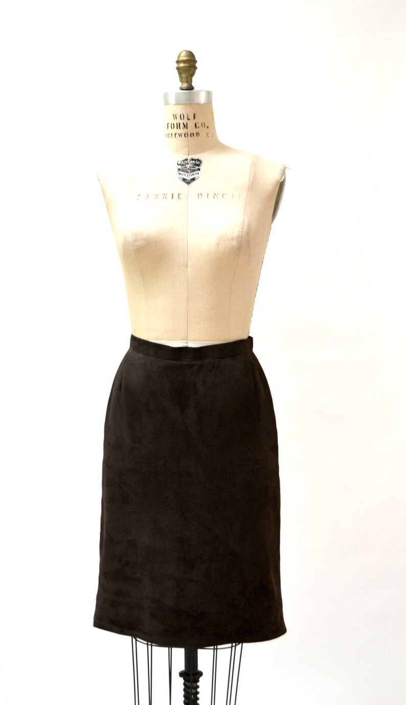 May include: A brown suede skirt on a dress form. The skirt is knee-length and has a straight silhouette.