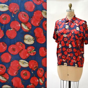 May include: A blue short-sleeved button-up shirt with a red apple print. The shirt has a collar and a button closure.