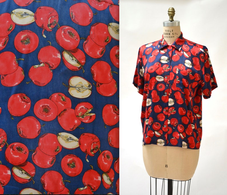 May include: A blue short-sleeved button-up shirt with a red apple print. The shirt has a collar and a button closure.