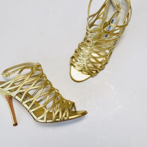 May include: A pair of gold-colored high-heeled sandals. The sandals feature a cage-like design with thin straps and a buckle closure. The heels are approximately 4 inches tall. The shoes are displayed on a white surface.