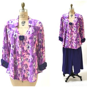 May include: A purple and pink floral print blouse with a blue collar and cuffs. The blouse has a V-neckline and a button closure. It is paired with a matching purple skirt.