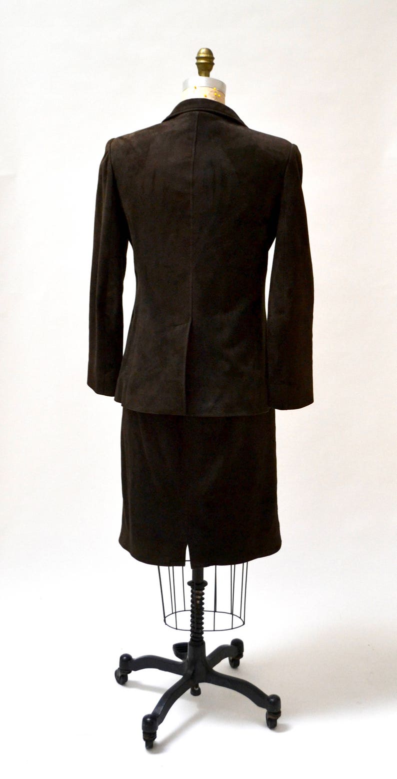 May include: A brown suede suit jacket and skirt set on a mannequin. The jacket has long sleeves and a classic cut. The skirt is knee-length and has a straight silhouette.