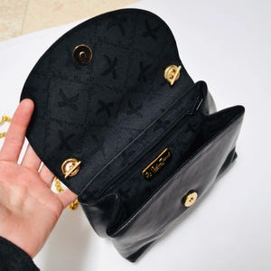 May include: Black leather handbag with a gold chain strap and a gold clasp. The inside of the bag is lined with black fabric and has a gold label that reads "Salvatore Ferragamo".