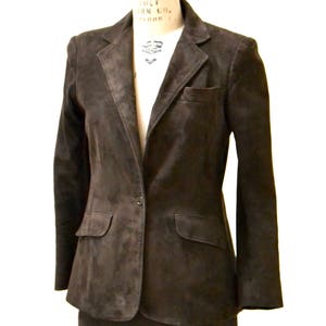 May include: A brown suede blazer and skirt suit. The blazer has a single button closure and two pockets. The skirt is knee-length.