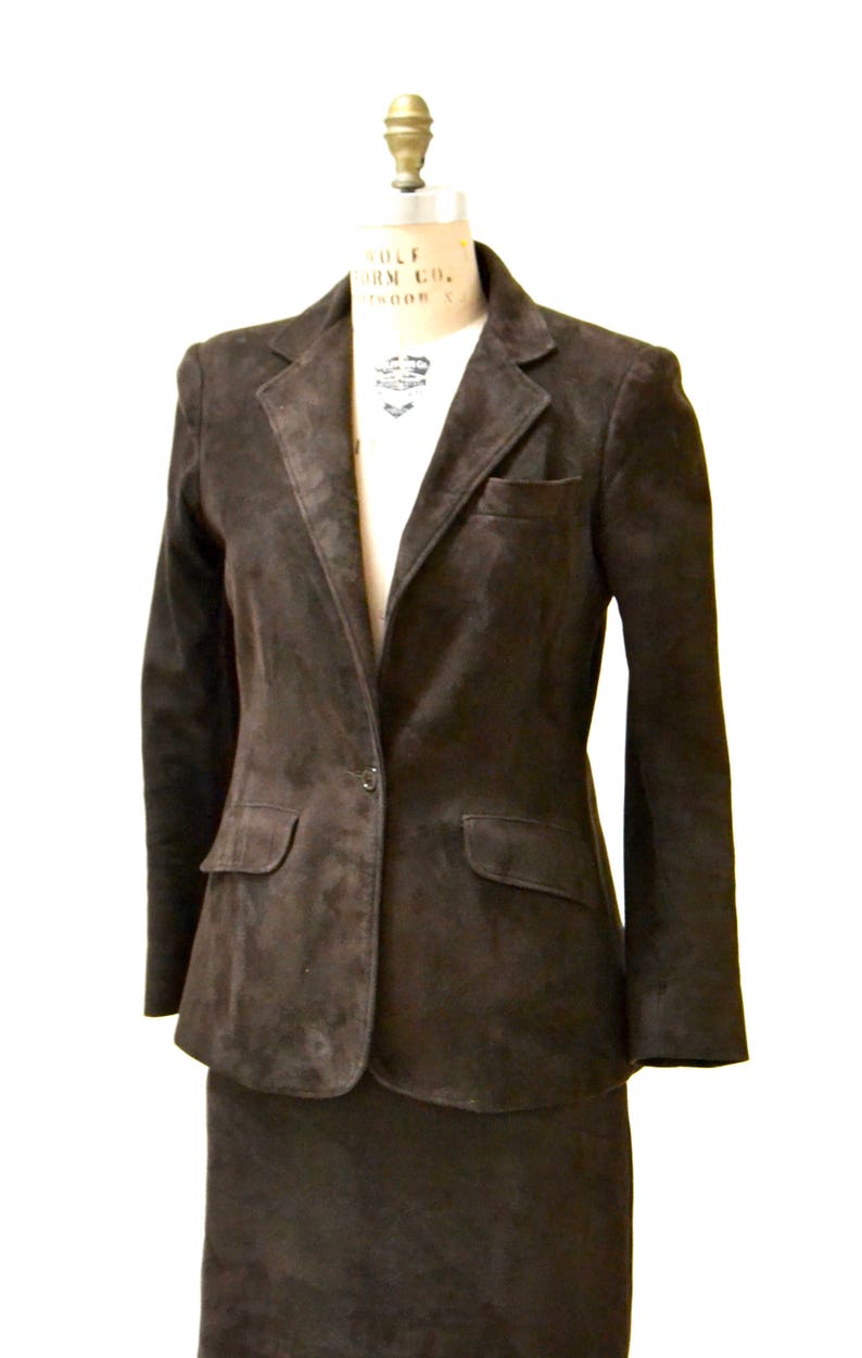 May include: A brown suede blazer and skirt suit. The blazer has a single button closure and two pockets. The skirt is knee-length.