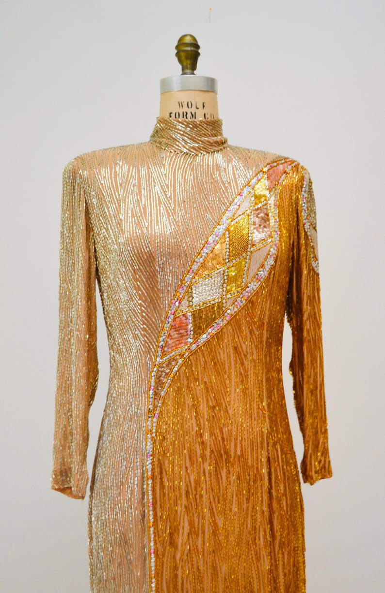 Op de afbeelding: Een lange, langarmige jurk met gouden en zilveren pailletten en een geometrisch patroon aan de rechterkant. De jurk heeft een hoge hals en een aansluitende pasvorm.