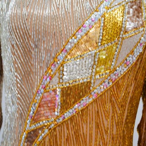Op de afbeelding: Een close-up van een kledingstuk van gouden en zilveren kralen. Het kledingstuk heeft een geometrisch patroon van gouden, zilveren en roze kralen.