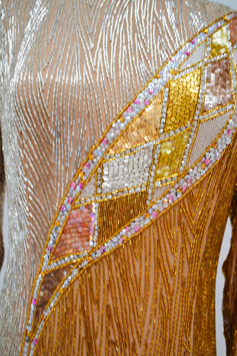 Op de afbeelding: Een close-up van een kledingstuk van gouden en zilveren kralen. Het kledingstuk heeft een geometrisch patroon van gouden, zilveren en roze kralen.