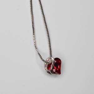 May include: A silver necklace with a pendant comprising two heart-shaped elements. One heart is a deep red gemstone, and the other is embellished with clear and iridescent crystals. The necklace is set against a plain white backdrop.