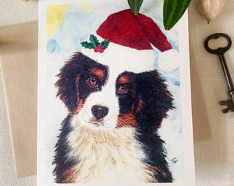 May include: A watercolor greeting card featuring a portrait of a Bernese Mountain Dog puppy wearing a red and white Santa hat with a small green holly sprig. The puppy has black, brown, and white fur, and the background is a soft blue wash.