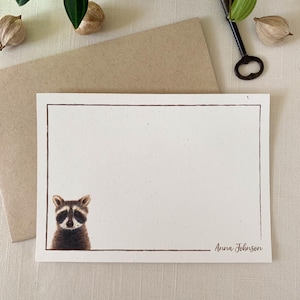 May include: A white card with a brown border and a raccoon illustration peeking from the bottom left corner. The text "Anna Johnson" is printed in brown at the bottom right corner.