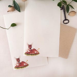 May include: Two off-white stationery sheets featuring a small watercolor illustration of a brown fawn resting in green grass with tiny yellow flowers. A dark metal skeleton key and green leaves are positioned near the paper edges.