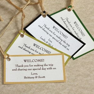 May include: Four welcome tags with different colored borders. The tags are made of paper and have a hole punched in the top. The text on the tags reads "Welcome! Thank you for making the trip and sharing our special day with us." The tags are tied with twine.