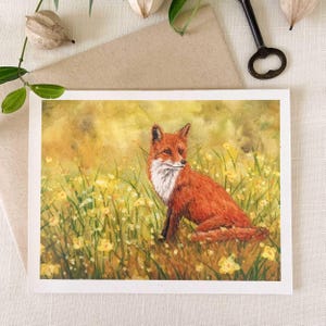 May include: A rectangular greeting card featuring a watercolor painting of a red fox with a white chest sitting in a field of yellow wildflowers and green grass. The fox is positioned in the center, looking toward the right side of the frame.