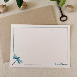 May include: A blank white card with a blue dragonfly illustration in the bottom left corner. The card has a blue border and the text "Anna Johnson" is written in blue ink in the bottom right corner.