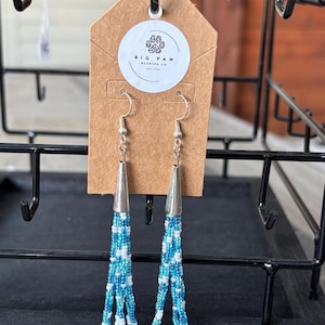 May include: A pair of beaded earrings with a silver cone top and multiple strands of blue and white beads. The earrings are displayed on a brown tag with a white circle that has a paw print logo and the text "BIG PAW BEADING CO."