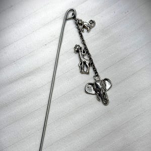 Puede incluir: Un marcapáginas plateado con tres colgantes: un mono, una jirafa y un elefante. Los colgantes están conectados por una delicada cadena y cuelgan de una varilla metálica larga y delgada. El marcapáginas está diseñado para libros.