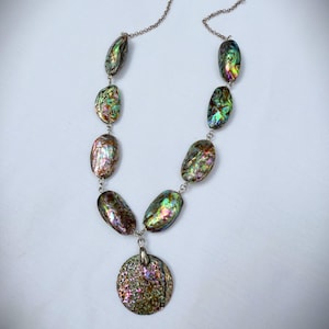 May include: Abalone shell necklace with iridescent colors of green, purple, and gold. The necklace features oval-shaped abalone shell beads connected by a silver chain, with a round pendant at the center. The necklace is a unique piece of jewelry.