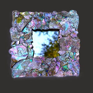 May include: A square mirror featuring a frame constructed from an assortment of iridescent abalone shell fragments. The shell pieces display a variety of colors including purple, blue, green, and pink, creating a textured, mosaic-like surface.