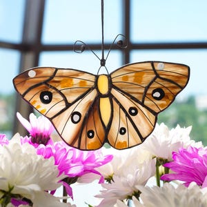 Puede incluir: Un atrapasoles de mariposa de vidrieras con detalles marrones, amarillos y negros. La mariposa está suspendida frente a una ventana, con flores blancas y rosas en primer plano. Las alas de la mariposa tienen un diseño.
