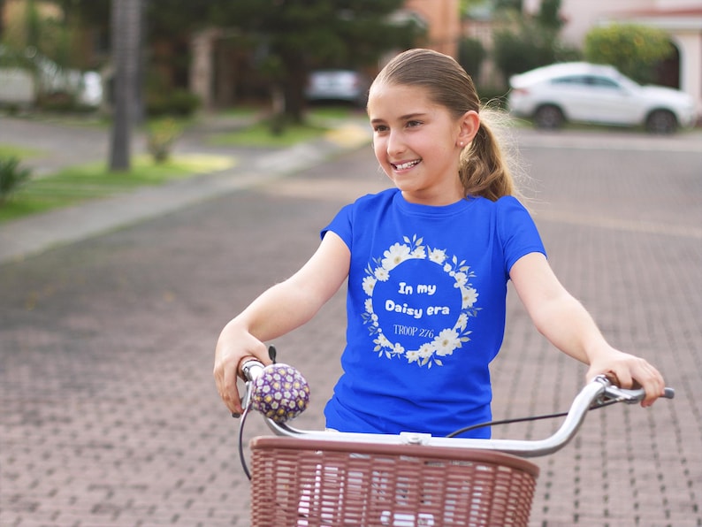 Op de afbeelding: Een jong persoon draagt een koningsblauw t-shirt met de tekst "In my Daisy era" in een bloemenkrans. Het shirt wordt gecombineerd met een fiets met een bruine mand en zilveren stuur. De fiets heeft een paarse en gele bel.