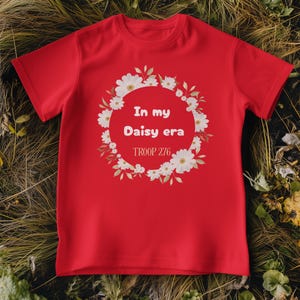 Op de afbeelding: Een rood t-shirt met de tekst "In my Daisy era" in een bloemenkrans. Het shirt bevat ook de tekst "TROOP 276" eronder. Het t-shirt wordt getoond op een bed van groene en bruine bladeren.