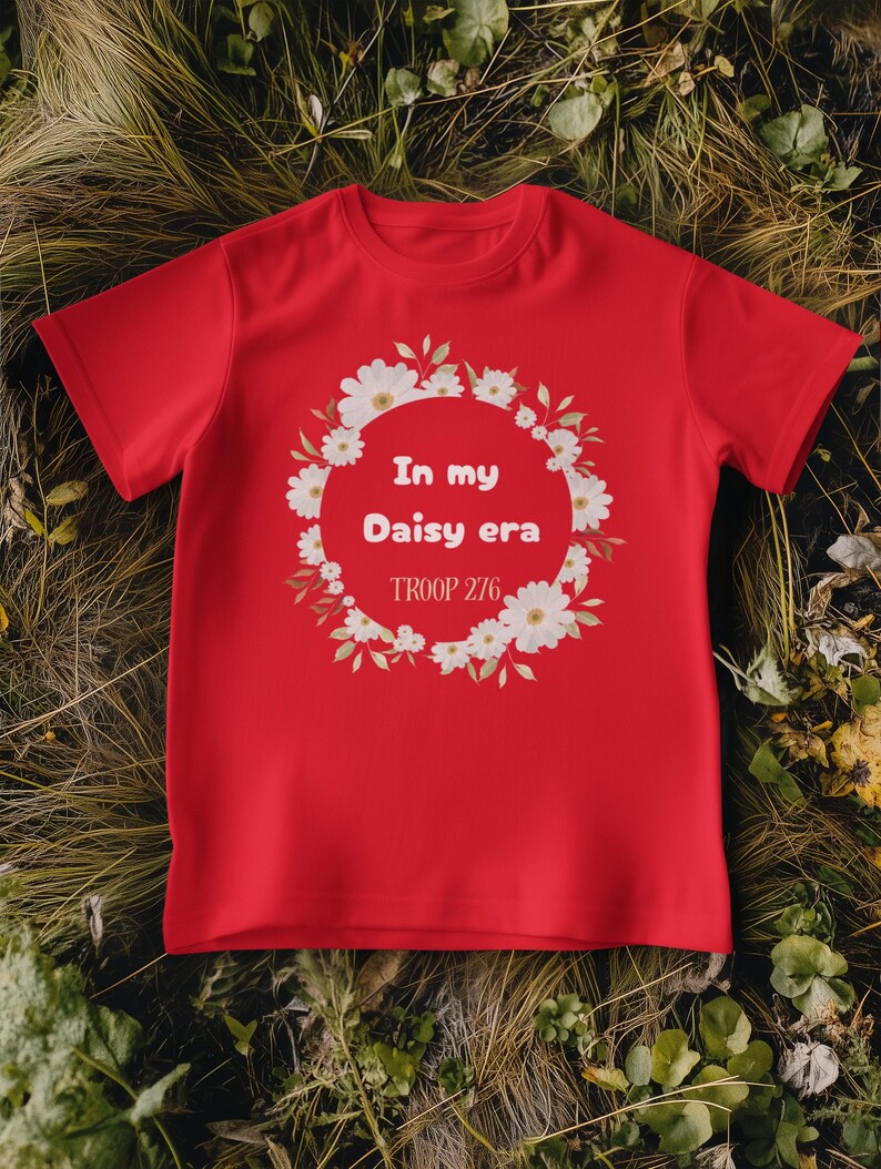 Op de afbeelding: Een rood t-shirt met de tekst "In my Daisy era" in een bloemenkrans. Het shirt bevat ook de tekst "TROOP 276" eronder. Het t-shirt wordt getoond op een bed van groene en bruine bladeren.