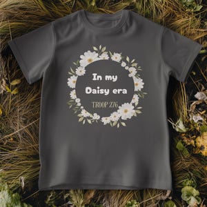 Op de afbeelding: Een donkergrijs t-shirt met een bloemenkransontwerp van witte madeliefjes en groene bladeren. De tekst "In my Daisy era" en "TROOP 276" staan in de krans. Het shirt wordt gepresenteerd op droog gras en bladeren.